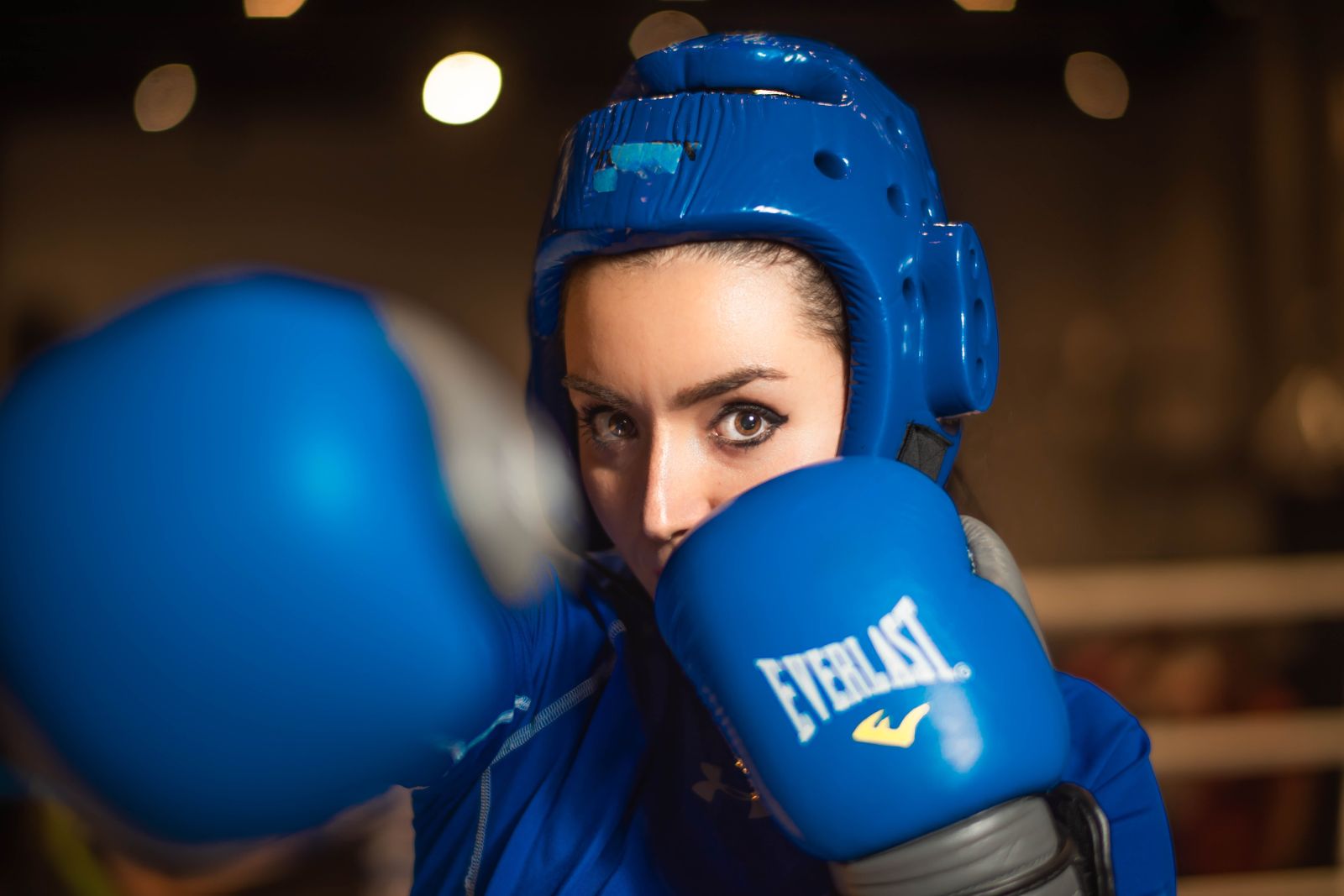 A female boxer wearing a blue headguard and gloves looks straight ahead as she throws a punch